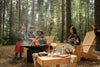 A family enjoys camping in the woods, with children roasting marshmallows over a campfire while parents relax on wooden chairs surrounded by tall pine trees—capturing the joy of outdoor adventures with kids | Kidspeek